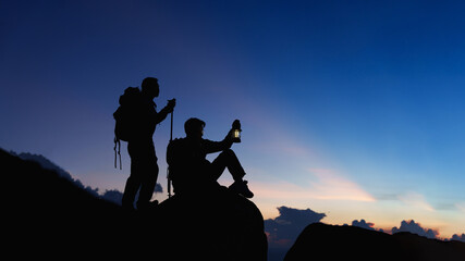 Silhouette of Asian two male standing raised hands with trekking poles and kerosene black lamp on...