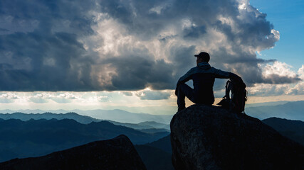Low angle view of Asian male hikers climbing mountain at sunset rays over the clouds with trekking poles sitting on cliff edge on top of rock mountain, Successful summit concept image.  