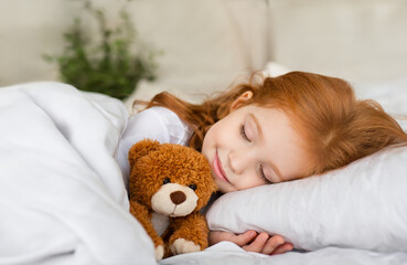 Cute little girl with red orange hair sleeping in bed with a teddy bear soft toy