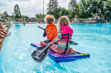 Two small kids paddling a cardboard boat in a race in a pool 