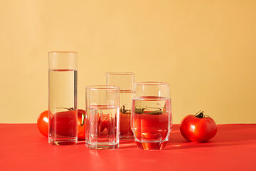 Transparent glasses of water are placed in front of juicy red tomatoes against a red and beige background. Tomatoes are vegetables that provide many necessary vitamins for the body.