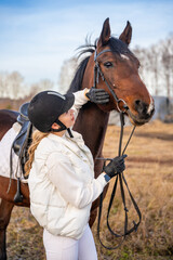 Obraz premium Pretty blond professional female jockey standing near brown horse. Friendship with horse