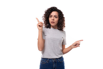 surprised european woman with black curly hair in a gray basic t-shirt points her fingers towards advertising space
