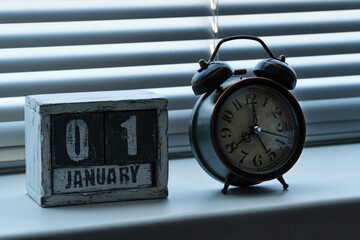 Morning of January 01 on wooden calendar standing window with blinds next to an alarm clock showing...