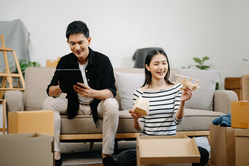 Asian young attractive couple man and woman use tablet and smartphone online shopping furniture decorate house with carton package move in new house. .