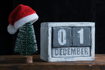 December 01 on wooden calendar standing next to Christmas tree decorated with santa claus hat.