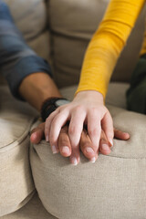 Diverse couple sitting on couch and holding hands in living room at home, copy space