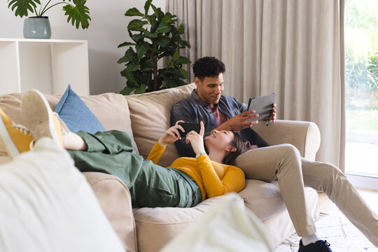 Happy diverse couple lying on couch, using tablet and smartphone in living room at home, copy space