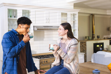 Happy diverse couple discussing and drinking tea in bright kitchen at home, copy space