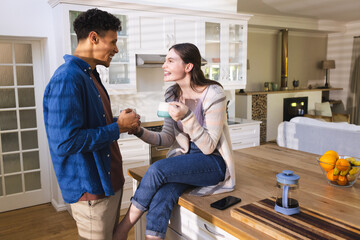 Happy diverse couple discussing, touching hand and drinking tea in kitchen at home, copy space