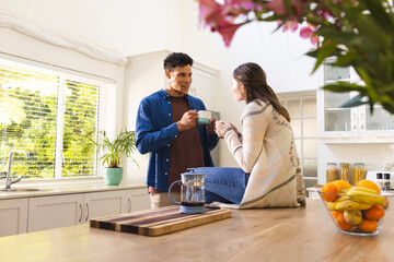 Happy diverse couple discussing and drinking tea in bright kitchen at home, copy space