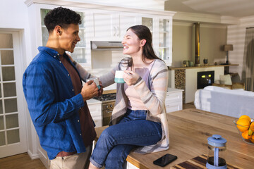 Happy diverse couple discussing, touching shoulder and drinking tea in kitchen at home, copy space