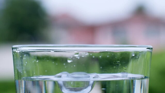 Droplets of water hitting the surface of crystal-clear water in a transparent glass cup on a blurry background.