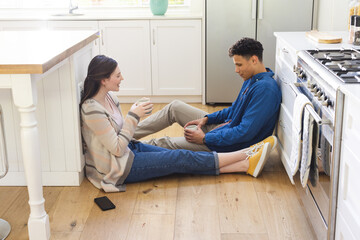 Happy diverse couple sitting on floor, discussing, holding tea cups in kitchen at home, copy space