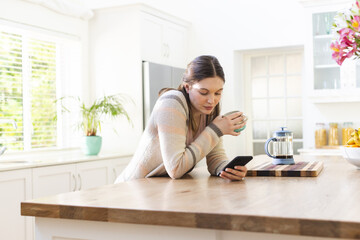 Focused caucasian woman holding mug and using smartphone in kitchen at home, copy space