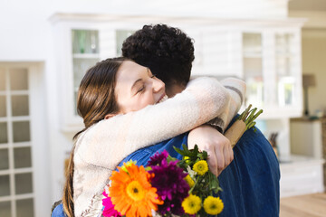 Happy diverse couple holding flowers and embracing in kitchen at home, copy space