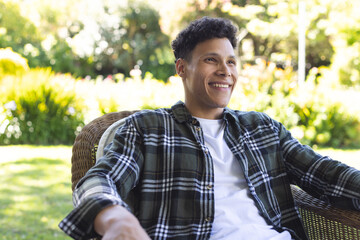 Portrait of happy biracial man sitting in chair in sunny garden, copy space