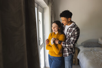 Happy diverse couple embracing and looking through window in bedroom at home, copy space