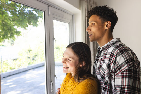 Happy diverse couple embracing and looking through window at home, copy space - Powered by Adobe