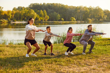 Family stretching after sport. Young family with two children is playing sports outside on bright sunny day. Mom, dad, son, daughter are squatting with outstretched arms against background of lake. - obrazy, fototapety, plakaty