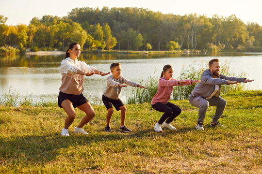 Family Stretching After Sport. Young Family With Two Children Is Playing Sports Outside On Bright Sunny Day. Mom, Dad, Son, Daughter Are Squatting With Outstretched Arms Against Background Of Lake.