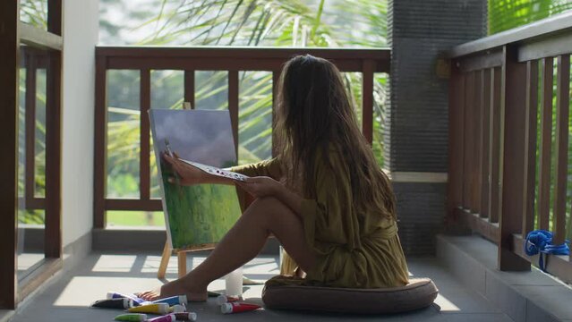 A young woman with brown hair paints with acrylic paints and her fingers on a canvas, the artist sits in a green robe on the floor and draws a picture on the canvas, the artist paints with her fingers