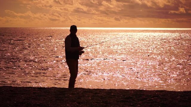 man standing on pier, holding a fishing rod and preparing to cast it into sea ocean at warm sunset. Man hobby fishing on sea tightens a fishing line reel of fish summer. Calm surface sea. Slow motion.