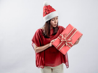 A portrait of a happy Asian woman celebrating Christmas, wearing a red dress and Santa hat, while carrying a gift box. Isolated against a white background.