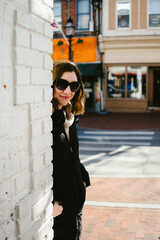 Fototapeta premium Woman in sunglasses leans against brick wall with urban buildings