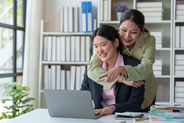 Two Asian office women cooperate and encourage each other sitting at desks with laptops. Discuss financial management planning Analysis of startup project ideas and office investment market statistics