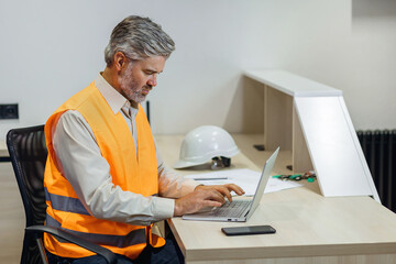 gray haired attractive construction bearded man in vest working with laptop, work on Home building project - obrazy, fototapety, plakaty
