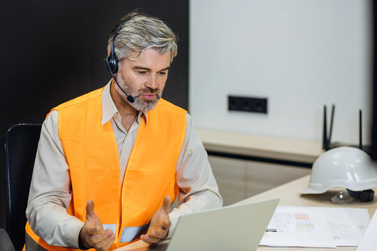 Old male architect sitting in the office consulting online using wireless headphones