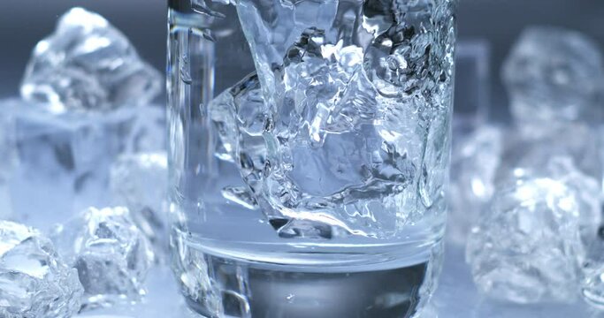 Super slow motion top view close up of ice cool cubes are falling with splashing water drops in transparent clear glass with pure fresh crystal liquid isolated on frozen background