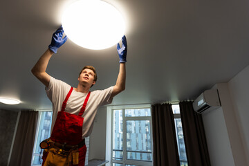 Worker installing lamp on stretch ceiling indoors.