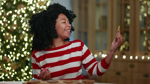 Middle-aged African American lady decorating Christmas cookies, looking, amiring the bakery, sitting at table with illuminated xmas tree in the background. High quality 4k footage