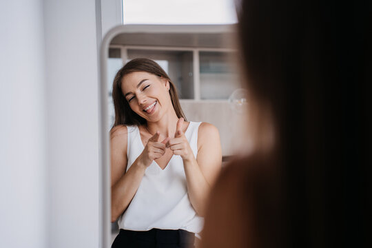 Cheerful young blonde girl smiling at mirror pointing at herself shows tongue being in playful mood ready for ;arty with friends. Careless female, freedom concept.