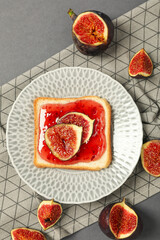 Piece of bread with jam and figs on plate on gray background, top view