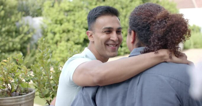 Happy diverse gay male couple embracing and smiling sitting in sunny garden, slow motion