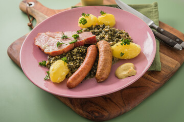 Oldenburger Grünkohl mit geräucherter Kohlwurst, Pinkel, Kassler Nacken, Kartoffeln, Senf, Petersilie auf Holzbrett, Serviette, Teller pink, und grün Hintergrund