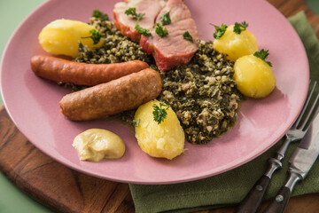 Oldenburger Grünkohl mit geräucherter Kohlwurst, Pinkel, Kassler Nacken, Kartoffeln, Senf, Petersilie auf Holzbrett, Serviette, Teller pink, und grün Hintergrund