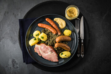 Oldenburger Grünkohl mit geräucherter Kohlwurst, Pinkel, Kassler Nacken, Kartoffeln, Senf, Petersilie auf Teller und dunkel schwarz Schiefer Hintergrund