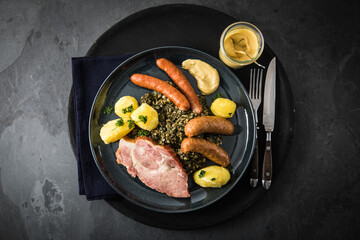 Oldenburger Grünkohl mit geräucherter Kohlwurst, Pinkel, Kassler Nacken, Kartoffeln, Senf, Petersilie auf Teller und dunkel schwarz Schiefer Hintergrund