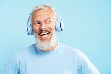 Portrait handsome smiling middle aged man, gray haired bearded  hipster listening music in wireless headphones isolated on blue background. Positive lifestyle, technology concept  
