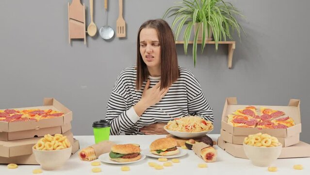 Sad unhealthy unhappy Caucasian woman wearing striped shirt sitting at table with big variety of fast food feeling stomachache and heartburn after eating junk food.