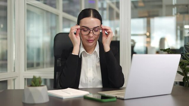 Ambitious female employee sitting and using modern devices while wearing glasses in bright room. Caucasian stylish brunette dressed in dark jacket smiling and putting hands on work dest indoor.