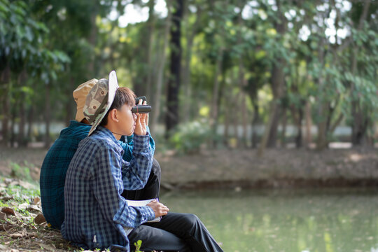 Asian boys sitting by the pond and holding binoculars to watch birds and learning nature and environment together.