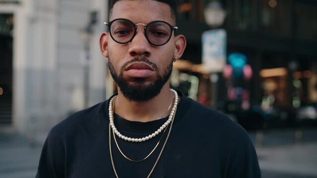 Close-up of young Black man with beard, wearing a black t-shirt and stylish glasses, adorned with chunky gold necklaces, set against a soft-focused urban background suggesting a lively city atmosphere