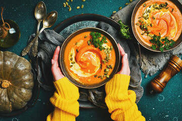 Female hands holding a bowl of pumpkin cream soup on a dark background. Autumn food concept. Top view.