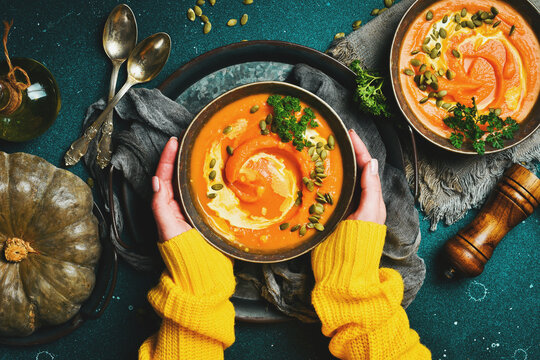 Female hands holding a bowl of pumpkin cream soup on a dark background. Autumn food concept. Top view.
