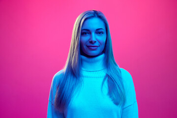 Close up portrait of young attractive smiling woman in knitted warm sweater against pink background in blue neon light.